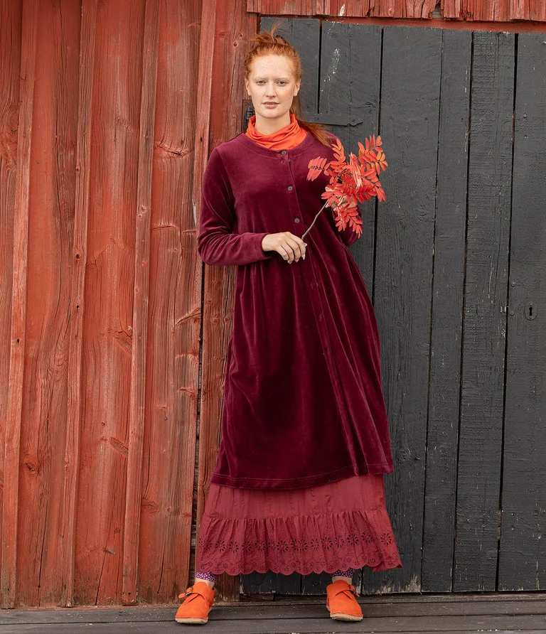 A woman standing up and wearing a fully buttoned red dress in velour from Gudrun Sjödén. She is holding a branch from a mountain ash tree in her hand.