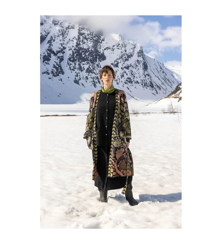 A woman standing in a snowy landscape wearing a patterned long cardigan over black clothes from Gudrun Sjödén’s 2025 Christmas collection.