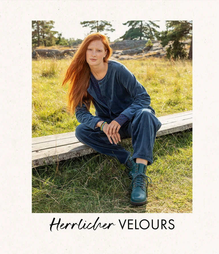 A woman sitting in a green meadow with the forest in the background, wearing blue velour clothing and blue boots from Gudrun Sjödén.