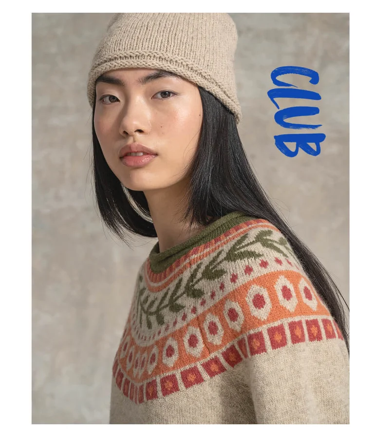 A woman wearing a natural-toned knitted wool sweater with a red, orange and green patterned yoke, and a knitted beanie from Gudrun Sjödén.