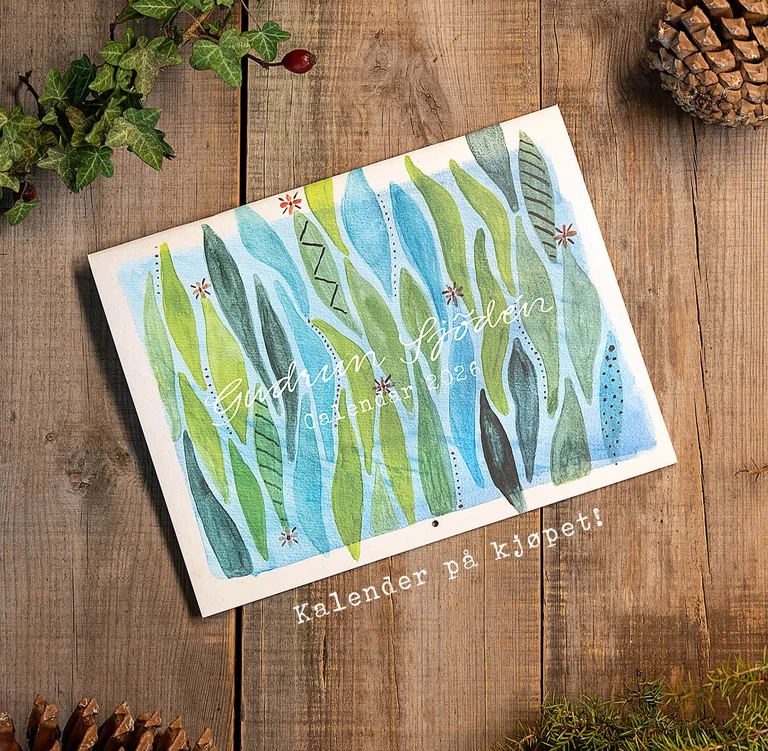 A calendar with watercolours from Gudrun Sjödén lying next to cones and greenery on a wooden surface. 