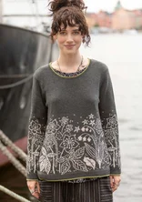 Eine Frau steht am Bootssteg mit dunkelgrauem Pullover. Der untere Teil des Oberteils ein helles Blumenmuster aufgestickt.