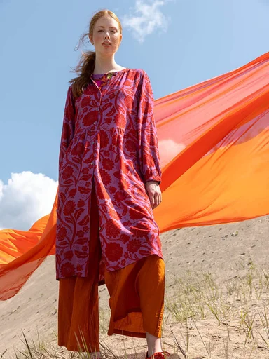 A woman standing on sand outdoors wearing layers of pink/red/orange clothes from Gudrun Sjödén. 