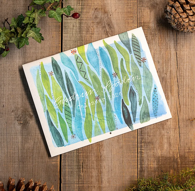 Een jaarkalender met aquarellen van Gudrun Sjödén op een houten ondergrond met dennenappels en groene takjes ernaast.