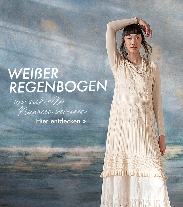 Eine Frau im weißen Lagenlook von Gudrun Sjödén, vor einem Hintergrund, der einem abstrakten Ölgemälde in blaugrauen Nuancen gleicht.