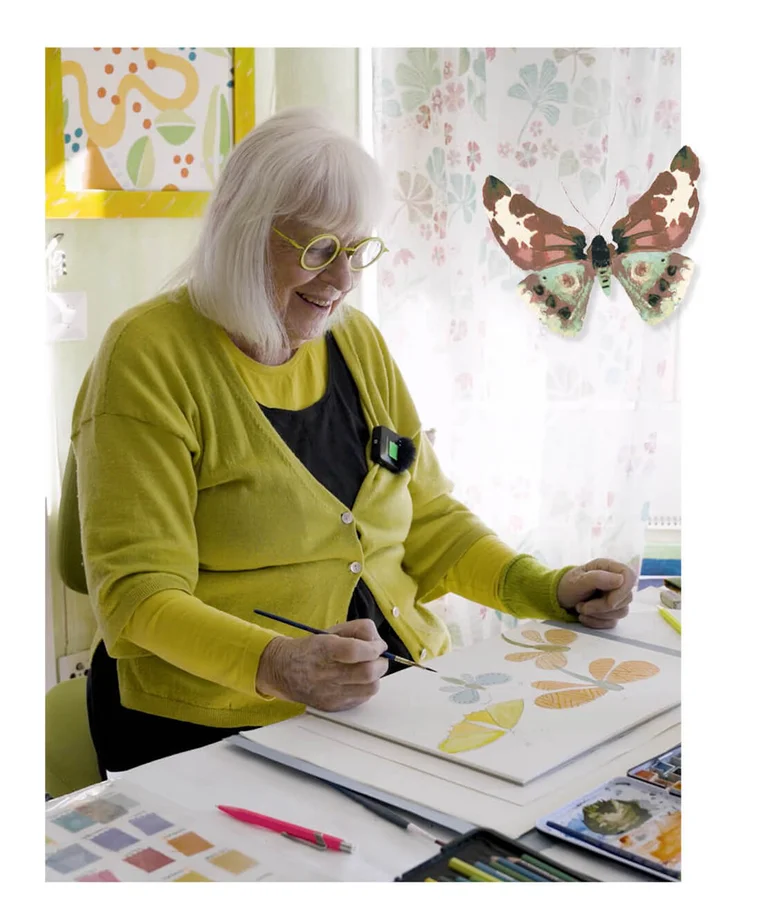 Gudrun Sjödén sitting at a desk painting watercolour butterflies. The picture also includes a standout image of a watercolour butterfly.