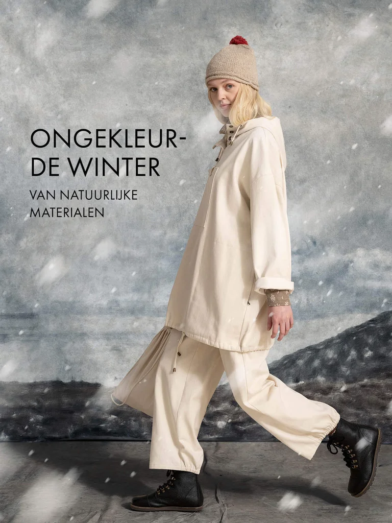 A woman walking while snow falls, wearing a knit hat, a white anorak and a pair of white pants from Gudrun Sjödén.