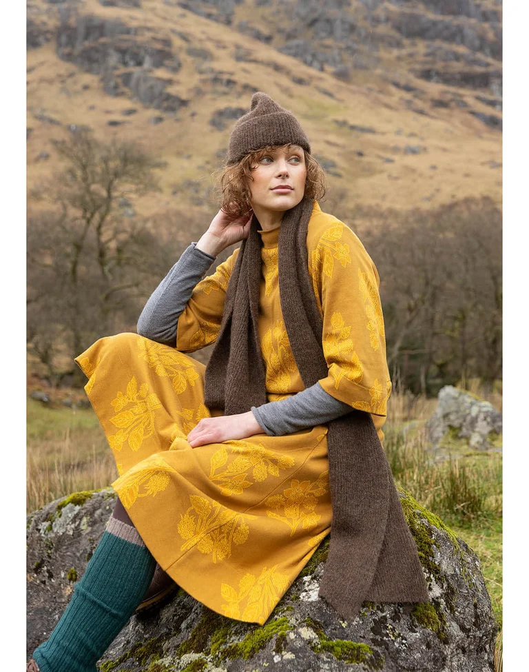A woman sitting outdoors wearing a yellow dress and brown, knitted accessories from Gudrun Sjödén.
