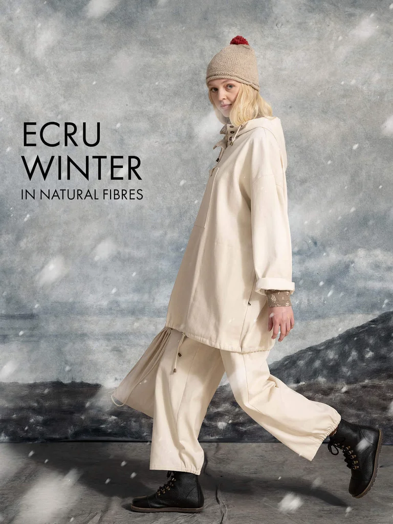 A woman walking while snow falls, wearing a knit hat, a white anorak and a pair of white pants from Gudrun Sjödén.