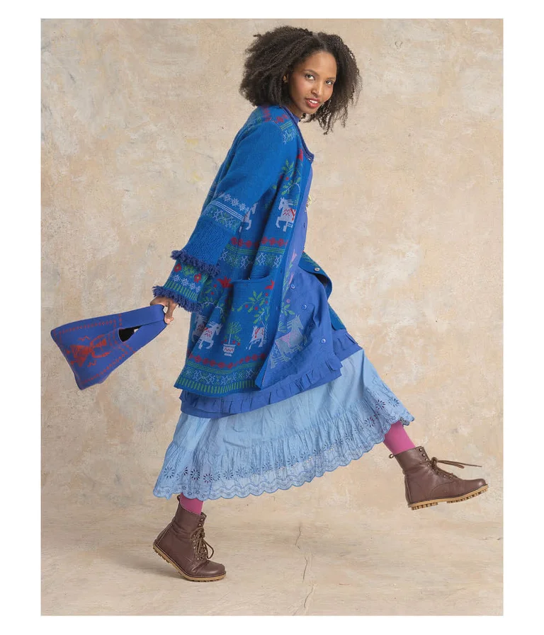 A woman mid-stride wearing a long blue jacquard-knit cardigan with fringed sleeves, a light-blue skirt and brown boots from Gudrun Sjödén. She is holding a knitted bag from Gudrun Sjödén.