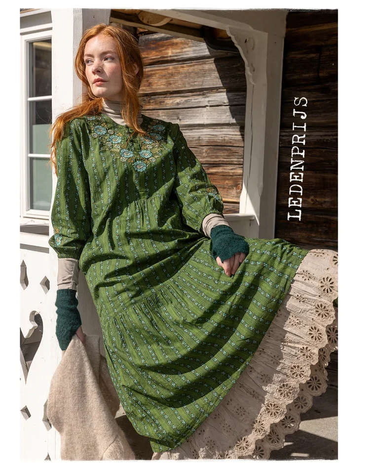 Een vrouw die op een brugreling zit en laagjes groene en beige kleding uit de kerstcollectie van 2025 van Gudrun Sjödén draagt.