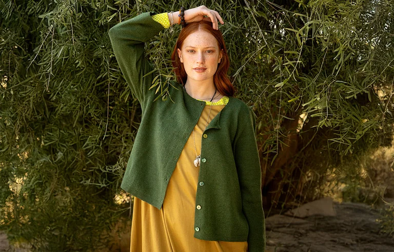 A woman standing next to a tree with one hand over her head, wearing a green knitted cardigan from Gudrun Sjödén’s Essentials collection AW 2025.