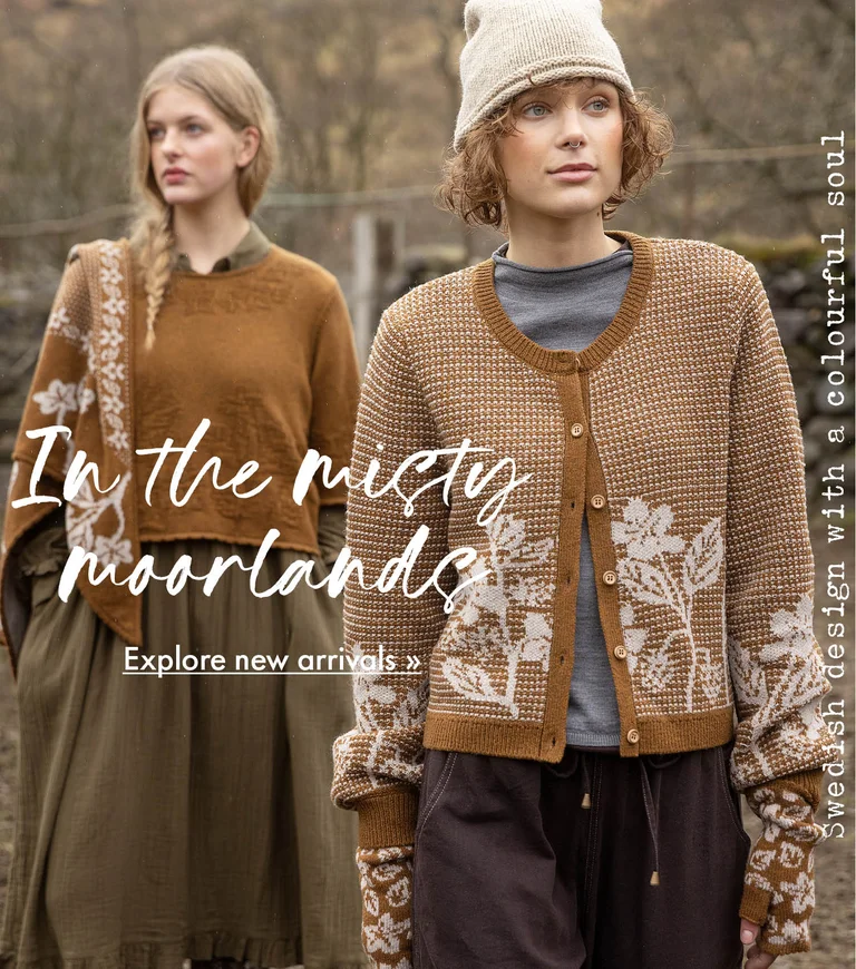 Two women standing next to each other wearing clothes in earthy hues from Gudrun Sjödén. Sheep and a stone wall are visible in the background.
