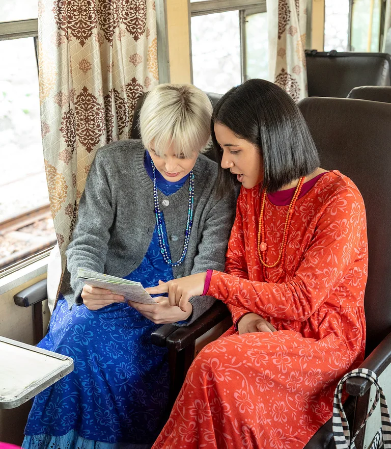 Two women sitting next to each other on a train wearing colorful clothing from Gudrun Sjödén. They are looking at a map.
