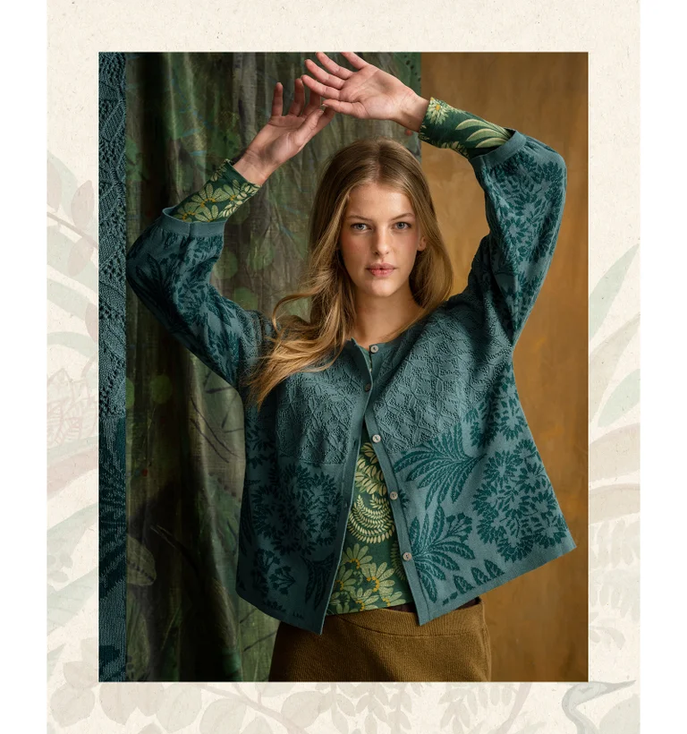 A woman with her hands over her head, wearing a patterned blue cardigan from Gudrun Sjödén.