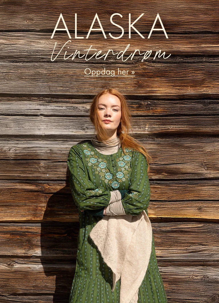 En kvinne som står mot en trevegg med armene i kryss kledd i en grønn kjole med broderier fra Gudrun Sjödéns julekolleksjon 2025. 