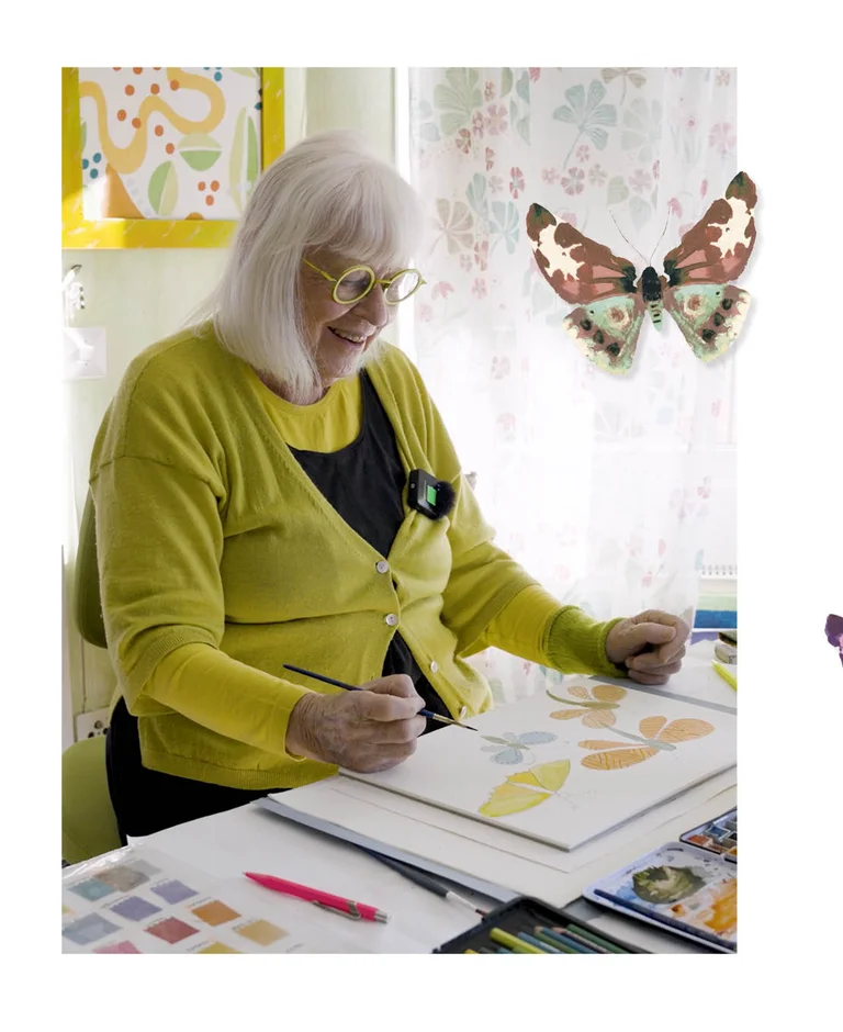 Gudrun Sjödén som sitter vid ett skrivbord och målar fjärilar med akvarellfärg. I bilden finns också en frilagd akvarellfjäril inlagd.