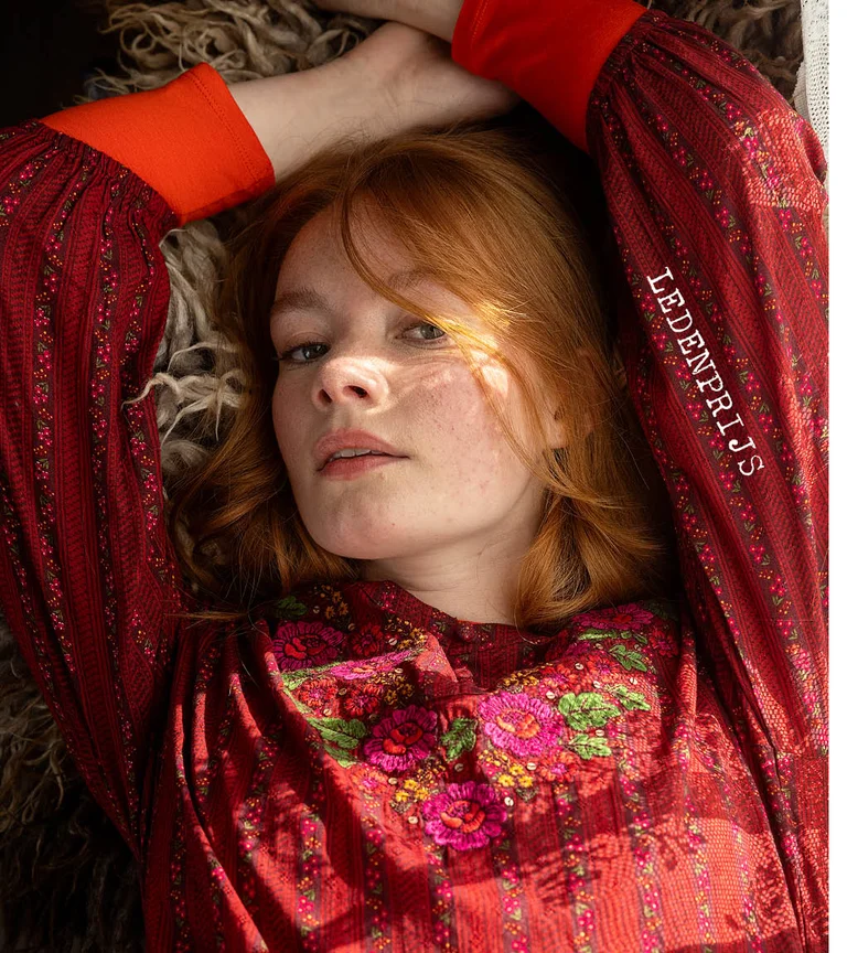 Een liggende vrouw in een rode, geborduurde jurk uit de kerstcollectie van 2025 van Gudrun Sjödén.