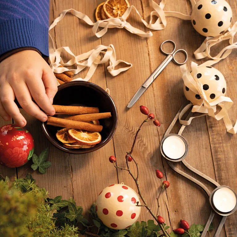 Handen die handgeschilderde kerstballen van Gudrun Sjödén versieren in een warme, creatieve omgeving met gedroogde sinaasappels en linten. 