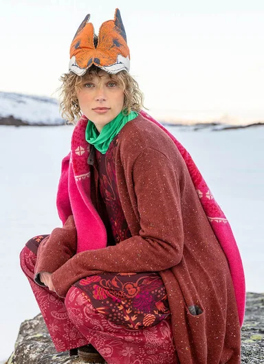 A woman with an animal mask on her head wearing a red and patterned outfit with a knitted cardigan from Gudrun Sjödén’s Alaska’s Wild Heart collection.