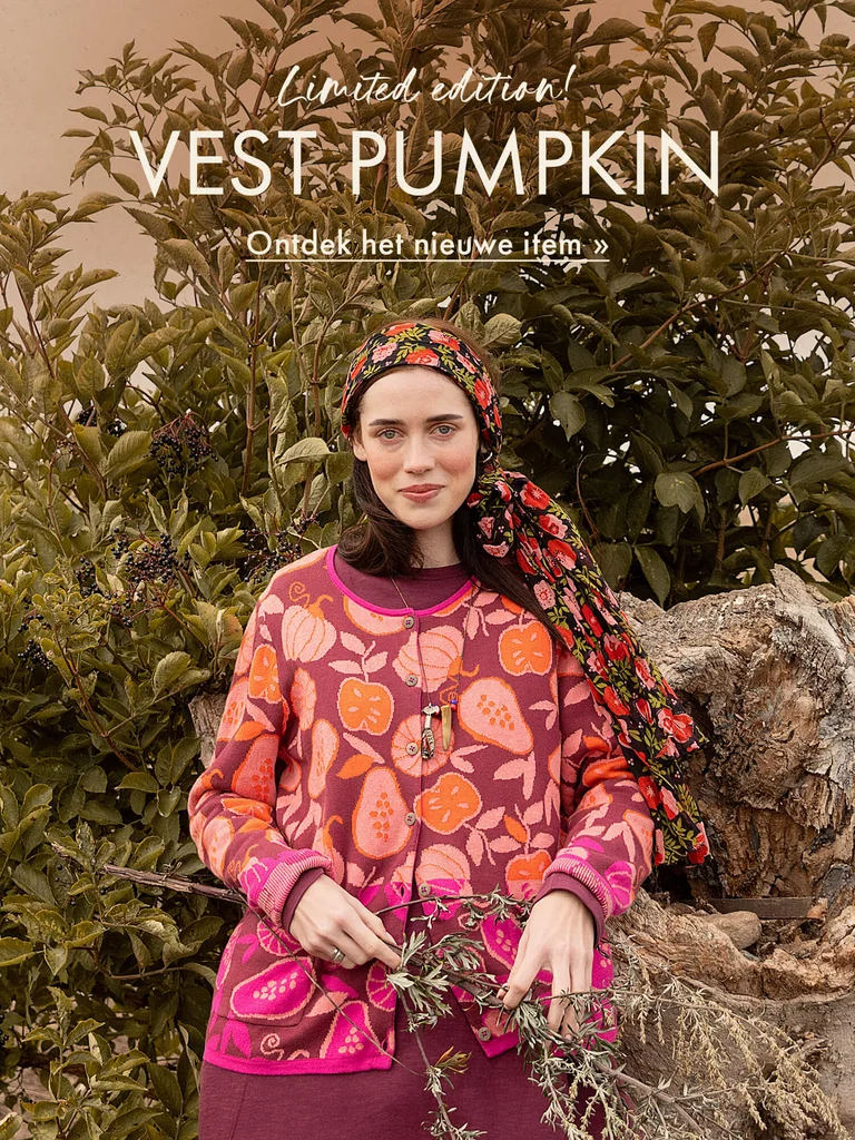 Een vrouw voor een groene struik in een herfstlandschap, gekleed in een gebreid vest van Gudrun Sjödén met een dessin van pompoenen en fruit in oranje en roze tinten.