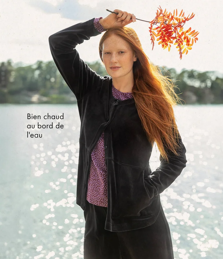 A woman in front of water wearing a black velour cardigan with matching velour pants from Gudrun Sjödén. She is holding a branch from a mountain ash tree in her hand.