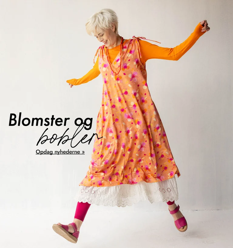 En kvinde som poserer med arme og ben udstrakt iført en orange, mønstret kjole over en orange top og et hvidt underskørt fra Gudrun Sjödén.