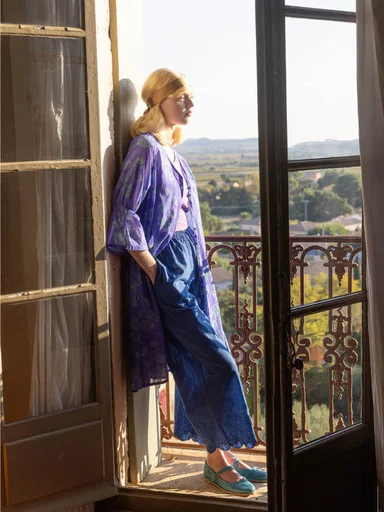 A woman wearing a light blue linen shirt from Gudrun Sjödén, standing by a fence, shading her eyes from the sun.