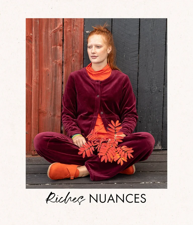A woman sitting outdoors wearing a red velour cardigan with matching velour pants from Gudrun Sjödén. She is holding a branch from a mountain ash tree in her hand.