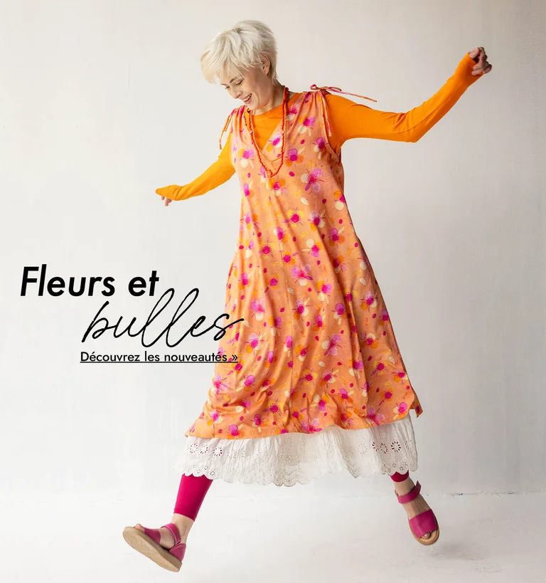 Une femme pose, bras et jambes tendus, elle porte une robe orange à motif sur un top orange et un jupon blanc signés Gudrun Sjödén.