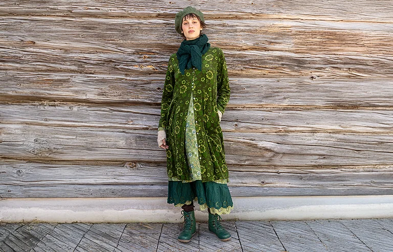 Une femme qui marche dans un paysage enneigé avec un masque animal et portant un pull à motif beige et gris provenant de la collection de Noël 2025 de Gudrun Sjödén. Sur la photo, on voit aussi un cœur graphique rouge.