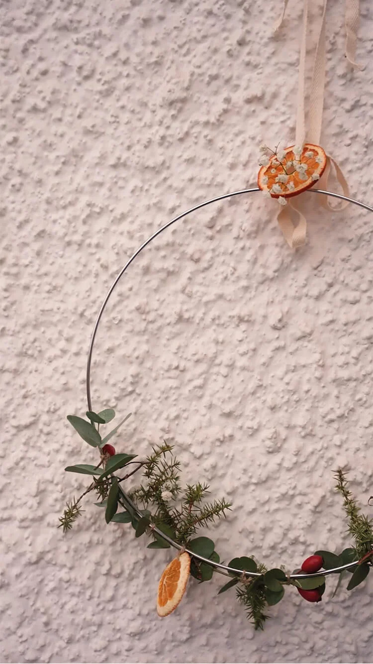 Handen versieren een kerstkrans op een metalen ring met groen, bessen en gedroogde sinaasappel.
