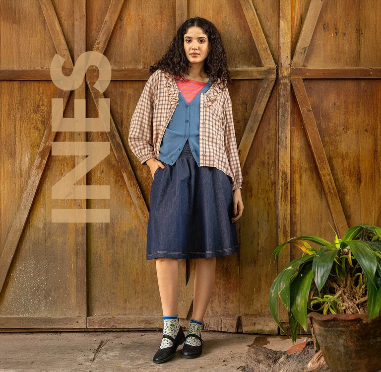 En kvinde som står lænet mod en trævæg iført lag på lag af tøj fra Gudrun Sjödén.