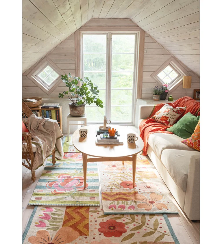 A loft with light wood panelling and Georgian windows. The floral mats on the floor and the red, orange and green cushions and throws on the sofa are from Gudrun Sjödén.