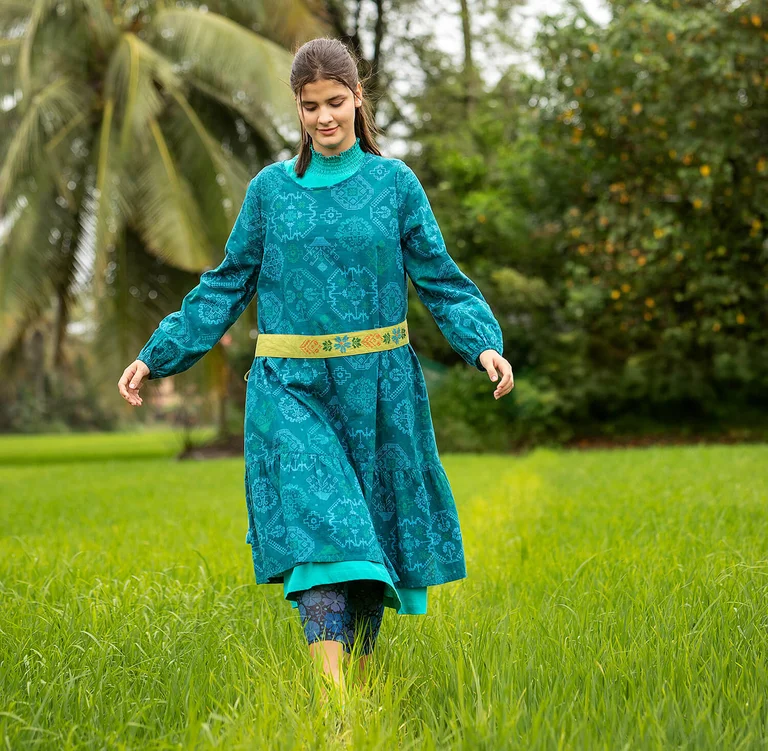 Une femme marchant sur une pelouse verte et portant une robe bleue à motif et une ceinture textile vert lime signées Gudrun Sjödén.