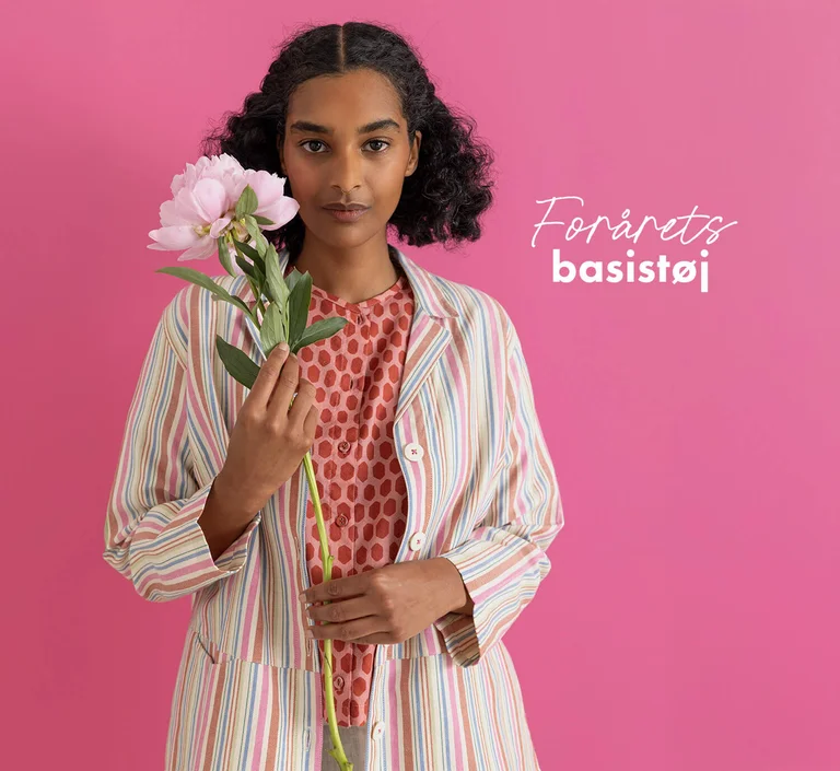 A woman standing against a pink background wearing essentials from Gudrun Sjödén’s 2026 Spring collection. Holding a pink flower in her hand.