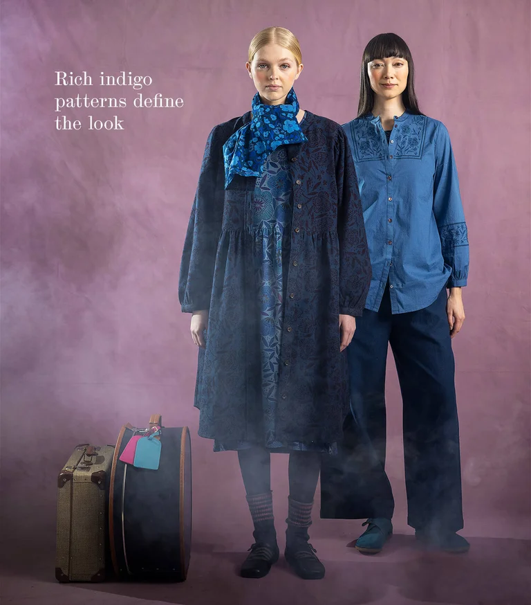 Two women next to each other wearing layers of clothing in shades of blue from Gudrun Sjödén. There are suitcases on the floor next to them.