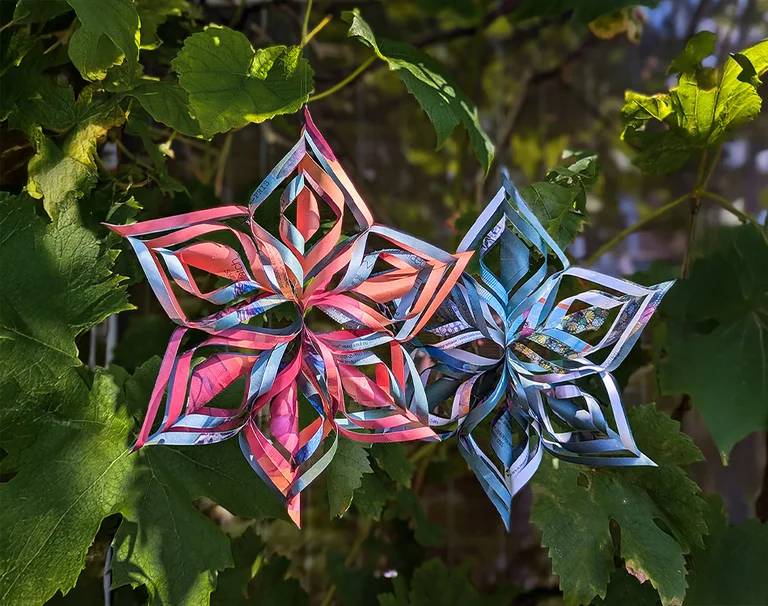 Zwei Papiersterne in Rosa/Lila/Blau, die an einem grünen Strauch hängen.