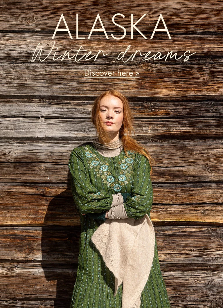 A woman standing against a timber wall with her arms crossed wearing a green dress with embroidery from Gudrun Sjödén’s 2025 Christmas collection. 