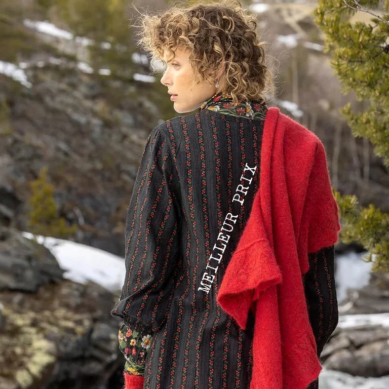 Une femme qui rit, allongée dans la neige, et portant le pull Luna à motif de hiboux provenant de la collection de Noël de Gudrun Sjödén. Sur la photo, on voit aussi un cœur graphique rouge. 
