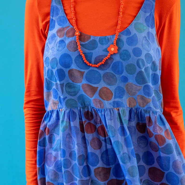 A close-up of the upper body of a woman wearing a red top under a blue, sleeveless dress from Gudrun Sjödén. 