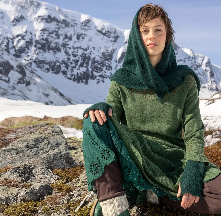 A woman sitting in front of a snowy mountain wearing layers of green clothes from Gudrun Sjödén’s 2025 Christmas collection.