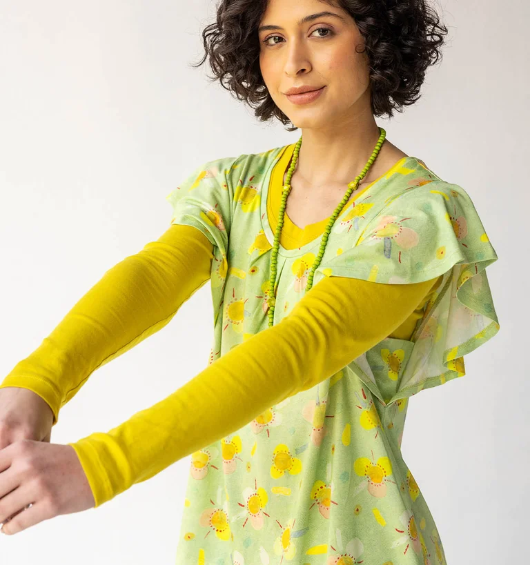 Une femme portant un top en jersey vert à motif sur un pull jaune à manches longues signés Gudrun Sjödén.