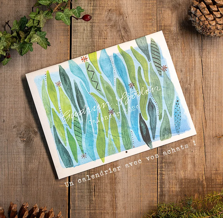 A calendar with watercolours from Gudrun Sjödén lying next to cones and greenery on a wooden surface.