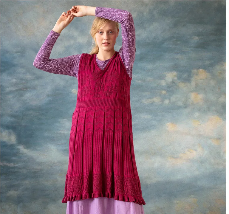 A woman holding one arm above her head, wearing a red knit dress over a light purple piece from Gudrun Sjödén.