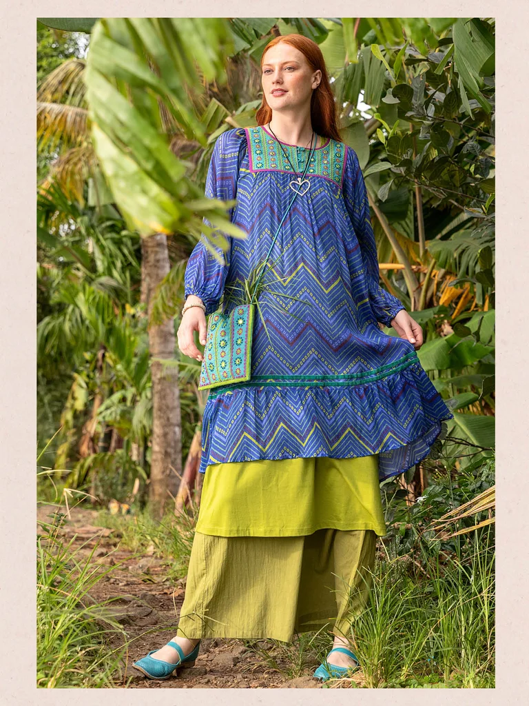 A woman standing in the jungle wearing layers of blue and green clothing from Gudrun Sjödén. 