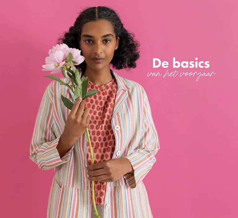 A woman standing against a pink background wearing essentials from Gudrun Sjödén’s 2026 Spring collection. Holding a pink flower in her hand.