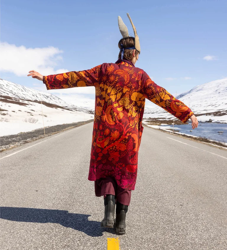 A woman standing on a road with her arms stretched out and her back to the camera. She is wearing a red, patterned long cardigan from Gudrun Sjödén’s 2025 Christmas collection.