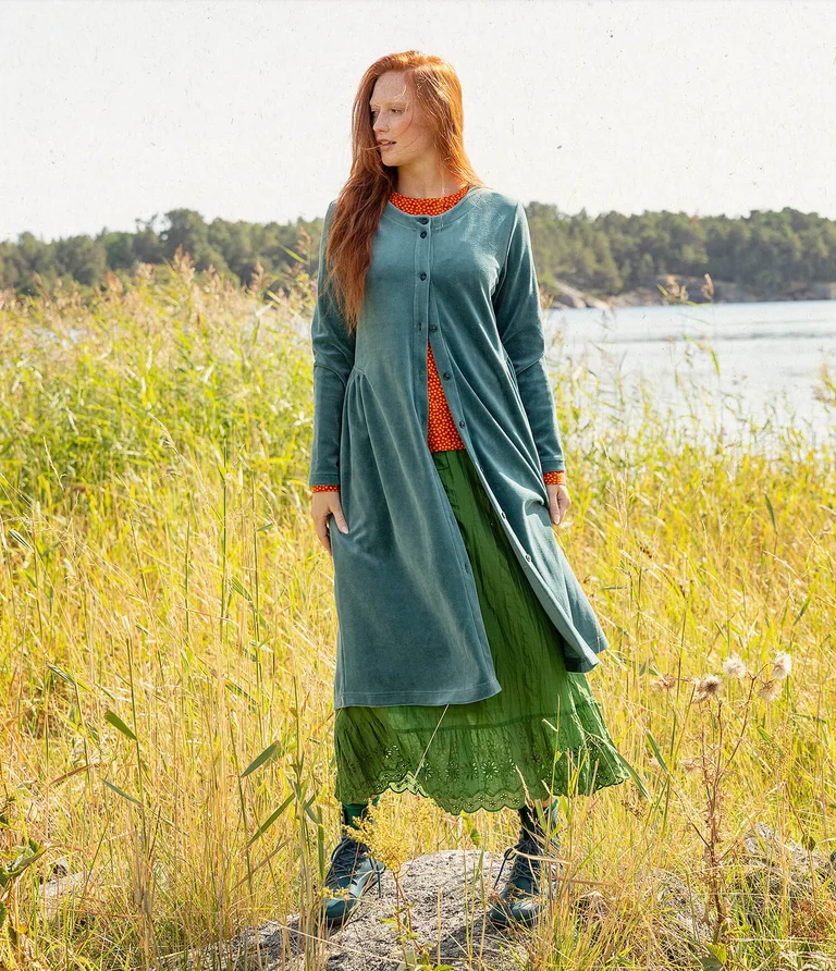 A woman standing in a green setting wearing a buttoned, turquoise velour dress from Gudrun Sjödén.