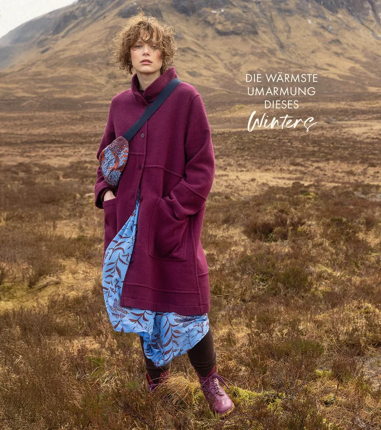 Eine Frau in einer Gebirgslandschaft in einem weinroten Wollmantel, einem hellblauen gemusterten Kleid und einer quer getragenen Bauchtasche von Gudrun Sjödén.